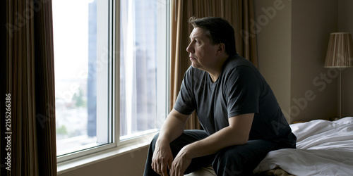 A man sits on the edge of a bed, gazing out a large window at a city skyline, lost in thought. He wears a dark t-shirt and trousers, his expression contemplative as he sits by the curtained window.