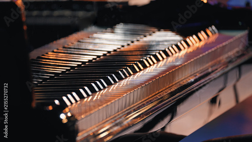 Closeup of piano keys illuminated by warm stage lights, shallow depth of field creates soft bokeh background. Polished black surface reflects golden glow