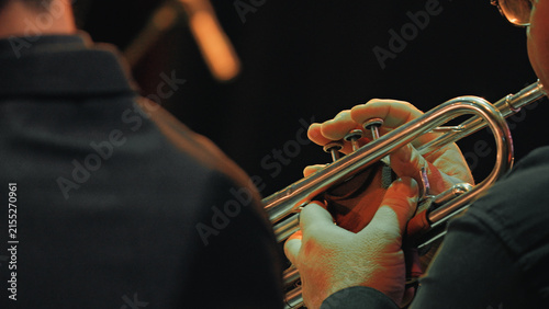 Closeup of musician hands playing trumpet under warm stage lights during symphony orchestra concert. Dark background highlights instrument fingers on valves. Evokes focus, precision, musical passion