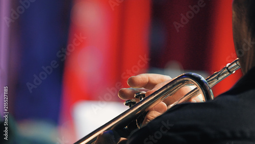 Closeup of musician playing trumpet during symphony orchestra concert. Hands on instrument, dark attire, vibrant red blue stage lights create dramatic mood. Ideal for music, performance, arts events