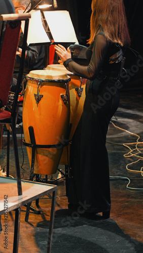 Female percussionist in black outfit playing conga drums under stage lights during symphony rehearsal. Warm orange drums contrast with dark stage