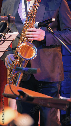Closeup of musician playing saxophone during live orchestra performance. Warm golden tones of instrument contrast with cool blue stage lighting. Formal suit, microphone, music stand visible