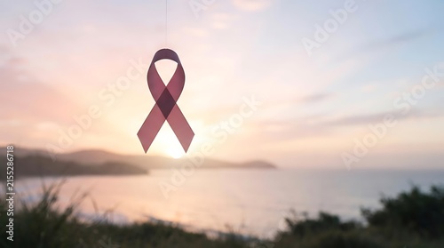 Delicate pink awareness ribbon hanging from thread against blurred coastal sunset landscape with ocean waves and green hills.