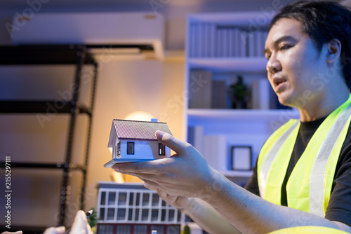 Two people are working together at a dimly lit desk. They are working on a home improvement project, the lighting adding to their focused expressions. Books and notebooks are stacked on shelves behind