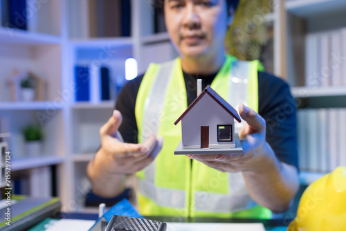 Two people are working together at a dimly lit desk. They are working on a home improvement project, the lighting adding to their focused expressions. Books and notebooks are stacked on shelves behind