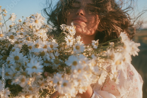 Woman surrounded by white daisies in a sunny meadow, with flowers framing her face and warm light creating a dreamy, natural summer portrait.
