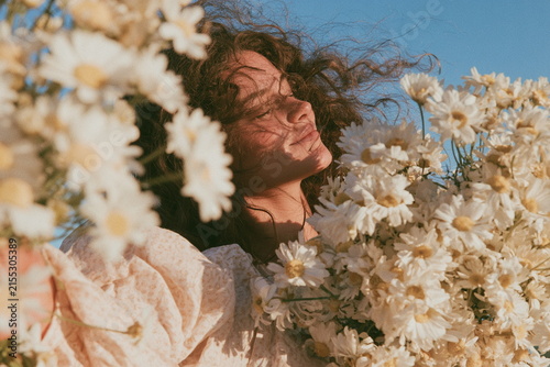 Woman surrounded by white daisies in a sunny meadow, with flowers framing her face and warm light creating a dreamy, natural summer portrait.