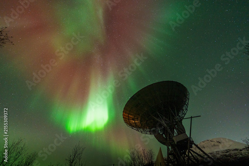 Ramfjordbotn, Troms, Norway - 07 October 2024: View of EISCAT Tromso radar dish illuminated by vibrant green and red aurora borealis in the night sky.