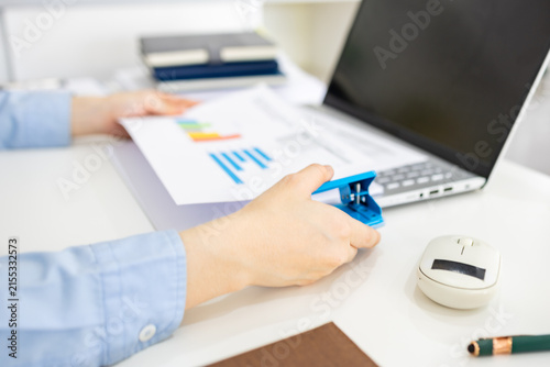 ands hold colorful charts beside a laptop. Fingers press a stapler, securing reports for analysis. Office desk holds a mouse, pen, and notebook. Focused work suggests data review or presentation prep
