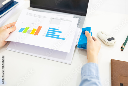ands hold colorful charts beside a laptop. Fingers press a stapler, securing reports for analysis. Office desk holds a mouse, pen, and notebook. Focused work suggests data review or presentation prep
