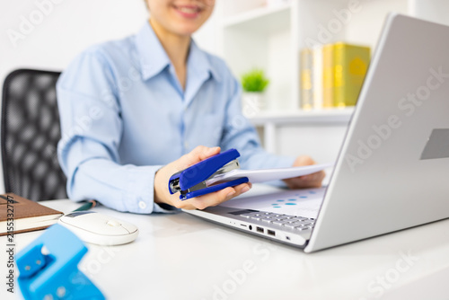 ands hold colorful charts beside a laptop. Fingers press a stapler, securing reports for analysis. Office desk holds a mouse, pen, and notebook. Focused work suggests data review or presentation prep

