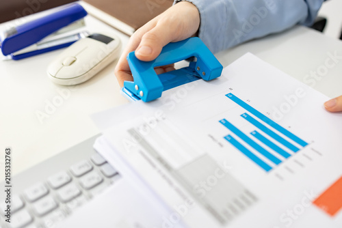 ands hold colorful charts beside a laptop. Fingers press a stapler, securing reports for analysis. Office desk holds a mouse, pen, and notebook. Focused work suggests data review or presentation prep
