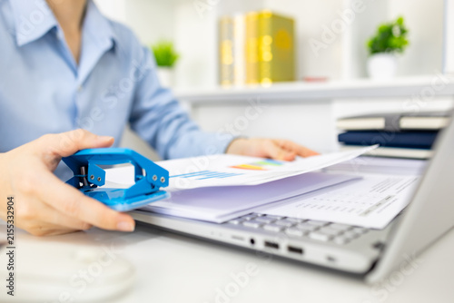 ands hold colorful charts beside a laptop. Fingers press a stapler, securing reports for analysis. Office desk holds a mouse, pen, and notebook. Focused work suggests data review or presentation prep
