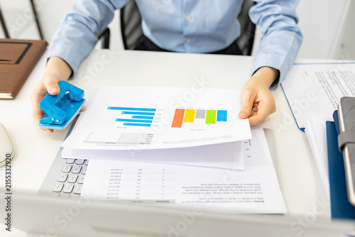 ands hold colorful charts beside a laptop. Fingers press a stapler, securing reports for analysis. Office desk holds a mouse, pen, and notebook. Focused work suggests data review or presentation prep
