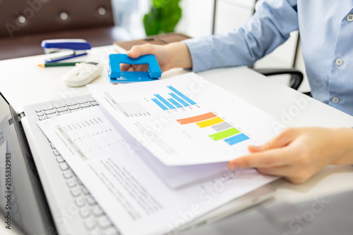 ands hold colorful charts beside a laptop. Fingers press a stapler, securing reports for analysis. Office desk holds a mouse, pen, and notebook. Focused work suggests data review or presentation prep
