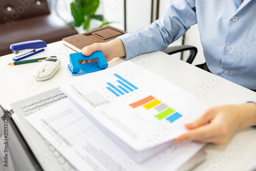 ands hold colorful charts beside a laptop. Fingers press a stapler, securing reports for analysis. Office desk holds a mouse, pen, and notebook. Focused work suggests data review or presentation prep

