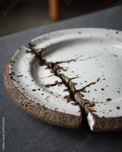 Broken ceramic plate with speckled white glaze and rough brown clay edges reveals deep crack as symbol of wabi sabi philosophy in minimalist setting.