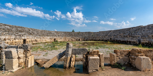 Ancient stone stadium ruins at Perge, a historic Roman archaeological site in Antalya, Turkey, featuring tiered seating and an open arena area under a sunny blue sky