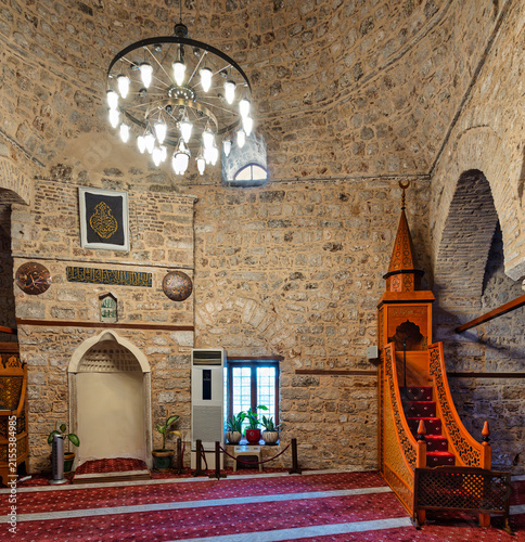 Interior view of the historic Yivli Minare Mosque featuring traditional stone walls, a wooden minbar, and an elegant chandelier in Antalya, Turkey