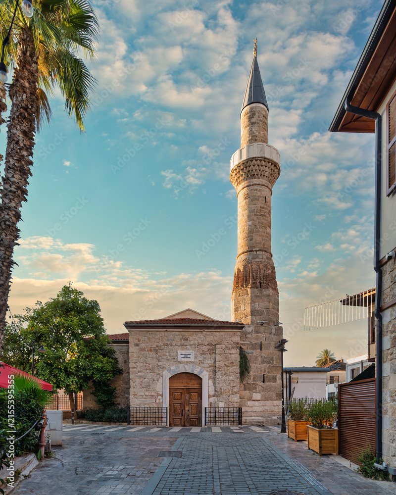 Fototapeta premium The historic Kesik Minare Mosque with its prominent minaret in the old town of Kaleici, Antalya, Turkey, showcasing stone architecture, palm trees, and a cobblestone street at sunset