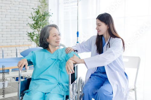 Asian female doctor consulting senior woman patient sitting in wheelchair in hospital clinic attentive comfort support quality elderly healthcare nursing assistance recovery wellness medical services.