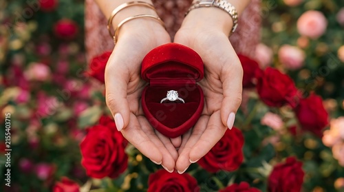 A person holding a heart shaped red velvet ring box with a diamond engagement ring inside surrounded by roses