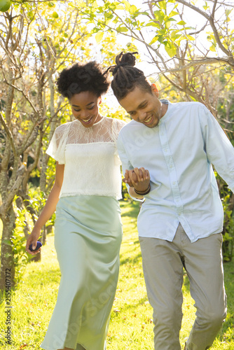 Canvastavla Couple walking holding hands in sunlit orchard, woman with smartphone, wearing w