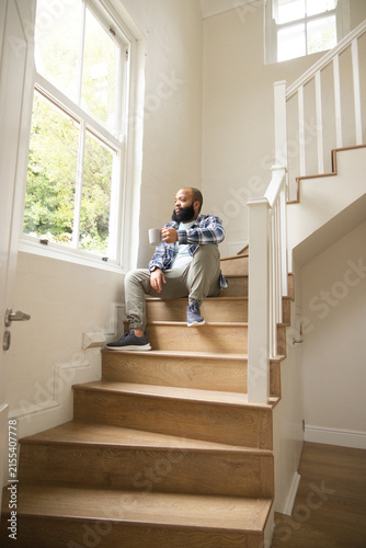 African American man sitting on wooden staircase at home holding ceramic mug and looking out window