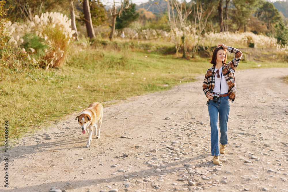 Fototapeta premium Woman walking her dog along a gravel path in a sunny countryside scene, casual attire, open landscape, and a peaceful outdoor moment of companionship and exercise.