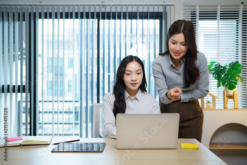 Two Happy Asian Businesswomen collaborating review work together at a modern office desk, discussing a project while reviewing information on a laptop in a bright professional workspace