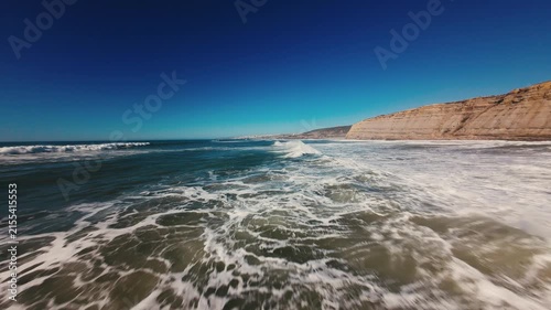 Waves crash over sand, foam swirls, sky glows deep blue. Rocks rise sharp against horizon, sea rushes with power. Bright day, wild beach, wind sings, water dances. Perfect for coastal scenes