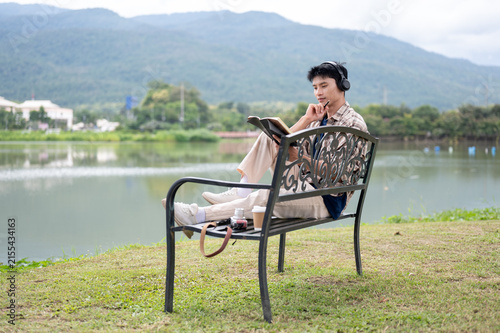 Asian Man Sitting on Park Bench Reading Book with Mountain and Lake View