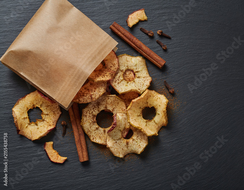 Delicious dried apple slices seasoned with warm cinnamon and fragrant cloves spill from a rustic brown paper bag onto a dark stone background, creating a comforting autumnal snack