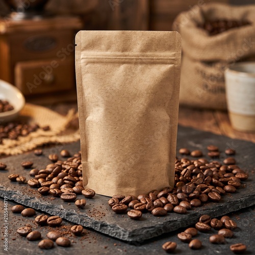 Natural unlabelled brown paper coffee bag packaging on slate stone surface with roasted coffee beans and vintage grinder, mockup for branding and design