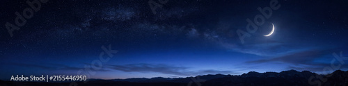 Panoramic view of a bright crescent moon in the deep blue night sky over a silhouetted mountain range