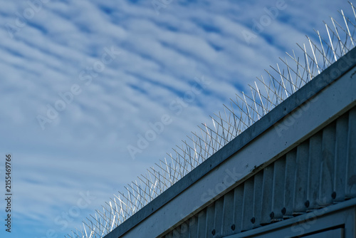 Konstfotografi Bird spikes run along a building roof's edge against a cloudy blue sky
