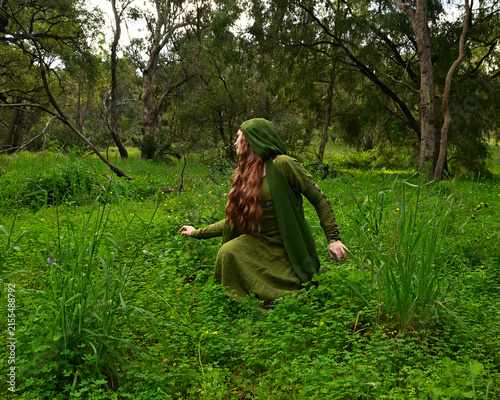 full length portrait of beautiful female model, sitting  kneeling figure picking herb plants, wearing hooded green medieval historical gown costume with cloak. Magical forest background with trees.
