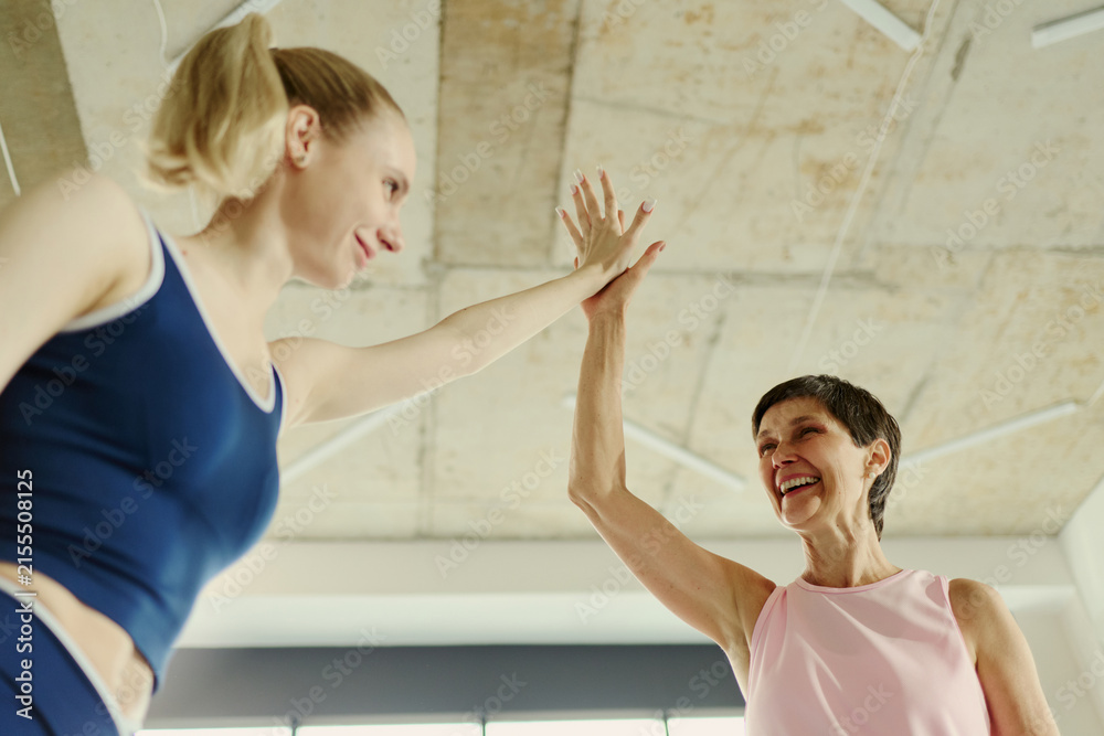 Fototapeta premium Young Caucasian woman and senior Caucasian woman giving high five in gym, celebrating fitness success. Low angle shot useful for active aging and intergenerational wellness campaigns