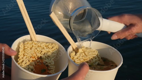 Two people preparing instant noodles outdoors, one holding two noodle containers while the other pours boiling water, hot meal for restoring energy after hiking, work and physical activity