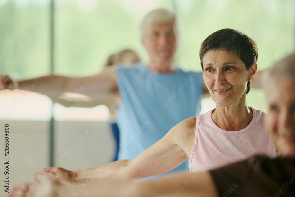 Fototapeta premium Senior Caucasian woman smiling during group fitness class in gym, promoting active aging and wellness. Image suitable for senior healthcare marketing and retirement lifestyle campaigns