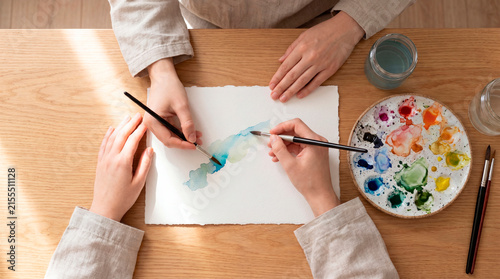 Top down view of two artists painting with blue watercolors on paper at a wooden desk. Ceramic palette, brushes, and colorful paints. Creative hobby and art therapy.