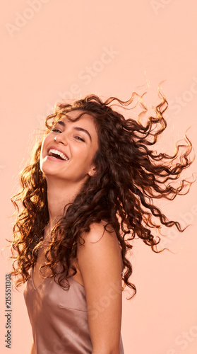 Happy beautiful young woman with flying curly dark hair laughing on peach pink background. Beauty and haircare concept.
