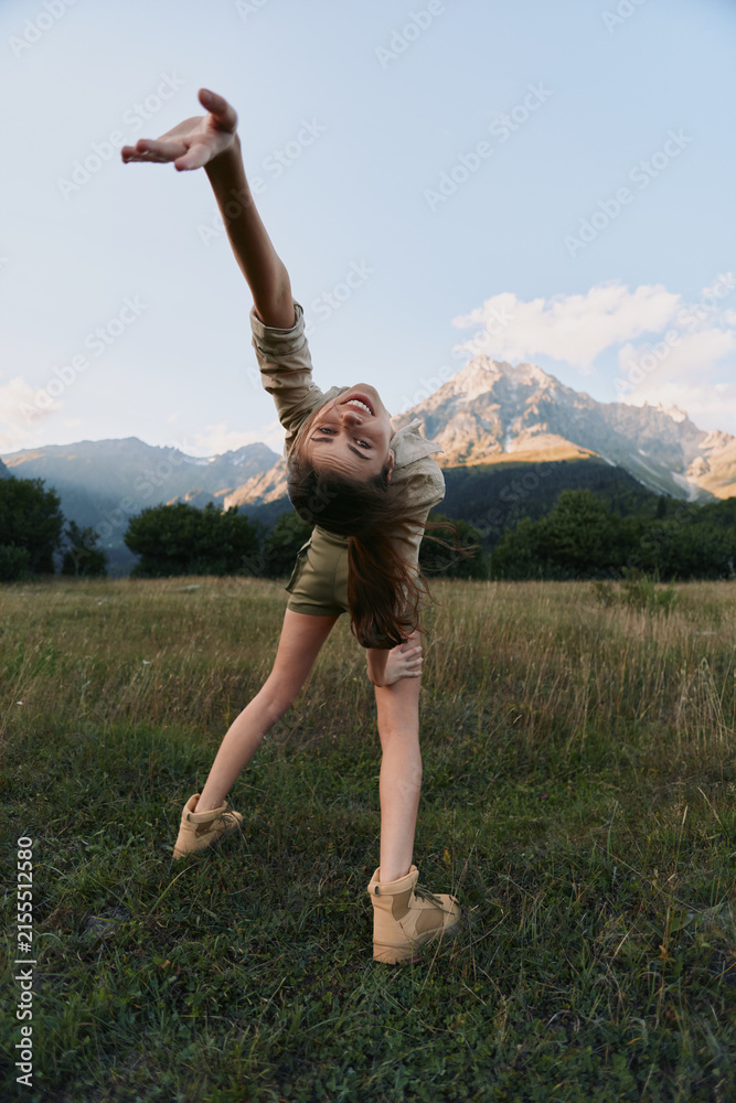 Obraz premium Young girl dances in a sunlit meadow with mountains in the background, arms raised in joy