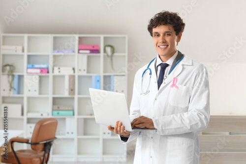Smiling male doctor wearing a pink ribbon and stethoscope, holding a laptop i...