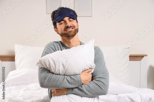 Handsome young man with sleep mask and pillow sitting in bed at home
