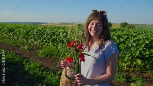 A happy young female teenager posing with bright red poppy flowers in a vast agricultural field under clear blue sky sunlight