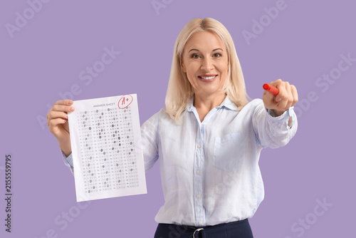 Mature female teacher with results of exam pointing at viewer on lilac backgr...