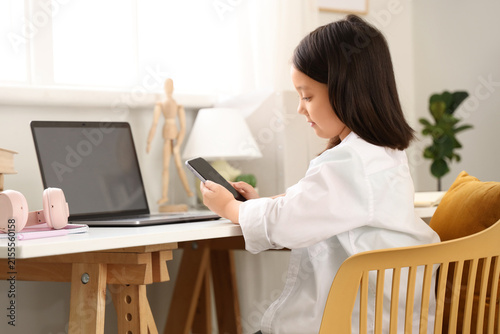 Little Asian girl with mobile phone and laptop studying online at home