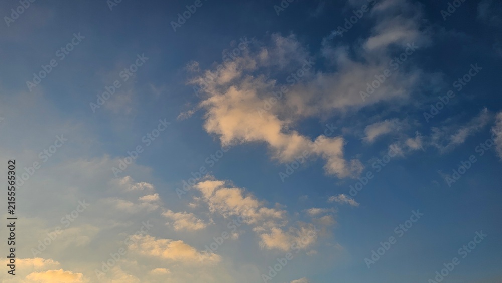 Fototapeta premium Fluffy white and golden-tinted cumulus clouds drift across a vivid blue sky during golden hour light.