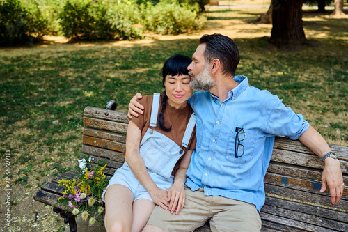 Foto Affectionate Couple Sitting on Park Bench With Flowers, Man Kissing Woman’s Chee